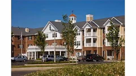 Three-story brick and siding senior living building with balconies, a covered entrance, parked cars, and landscaped grounds under a blue sky.