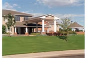 Exterior view of a senior living facility building with a covered entrance, well-maintained green lawn, trees, and a gazebo in the background under a partly cloudy sky.