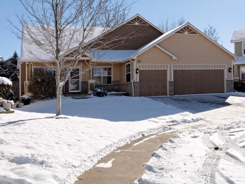 Single-story house with a three-car garage, snow-covered lawn and driveway, and a bare tree in front.