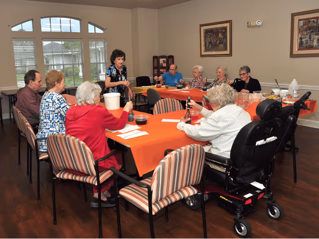 A group of elderly individuals seated around a large table with an orange tablecloth in a well-lit room. One woman stands at the table engaging with the group, who appear to be participating in an activity involving small containers and tools. The room has large windows with white blinds and framed artwork on the walls.