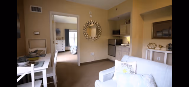 Interior view of a senior living facility apartment showing a small dining area with a table and chairs, a kitchenette with appliances, and a living area with a white couch. A doorway leads to a bedroom with a bed and dresser visible. The walls are painted beige and there is a decorative round mirror on the wall.