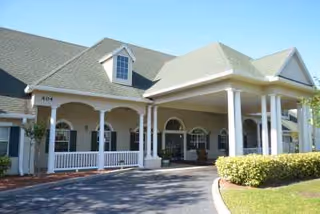 Front entrance of a residential senior living building with a covered porte-cochère, white columns, a wraparound porch, and landscaped driveway.
