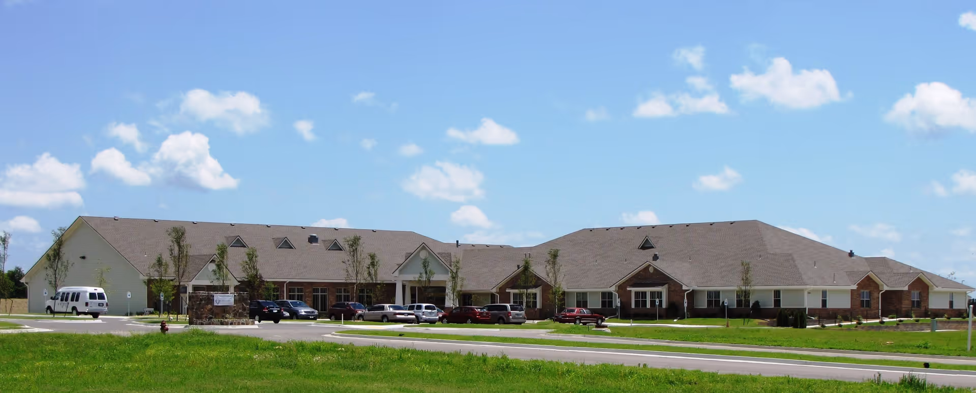 Wide exterior view of a single-story senior living facility building with a large parking area in front, surrounded by green grass and under a blue sky with scattered clouds.
