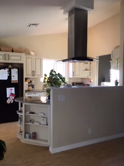 Open kitchen with a curved granite-topped island, stainless range hood, white cabinets, a black refrigerator, and a potted plant.