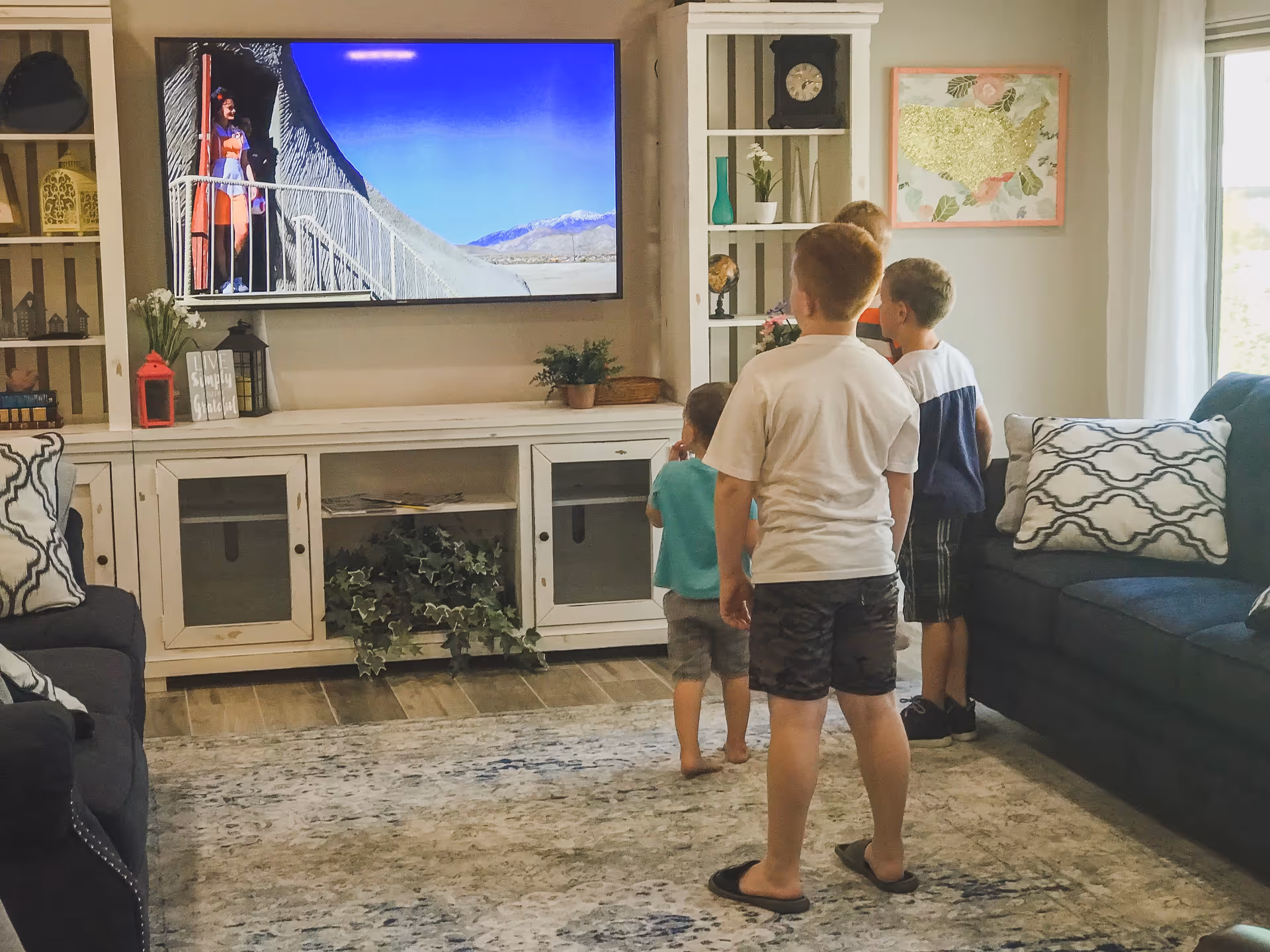A living room with four young boys standing and watching a large flat-screen TV mounted on the wall. The room has two dark blue sofas with patterned pillows, a white entertainment center with decorative items, and a light-colored rug on a wooden floor. There is a window with curtains on the right side and a framed floral artwork on the wall.