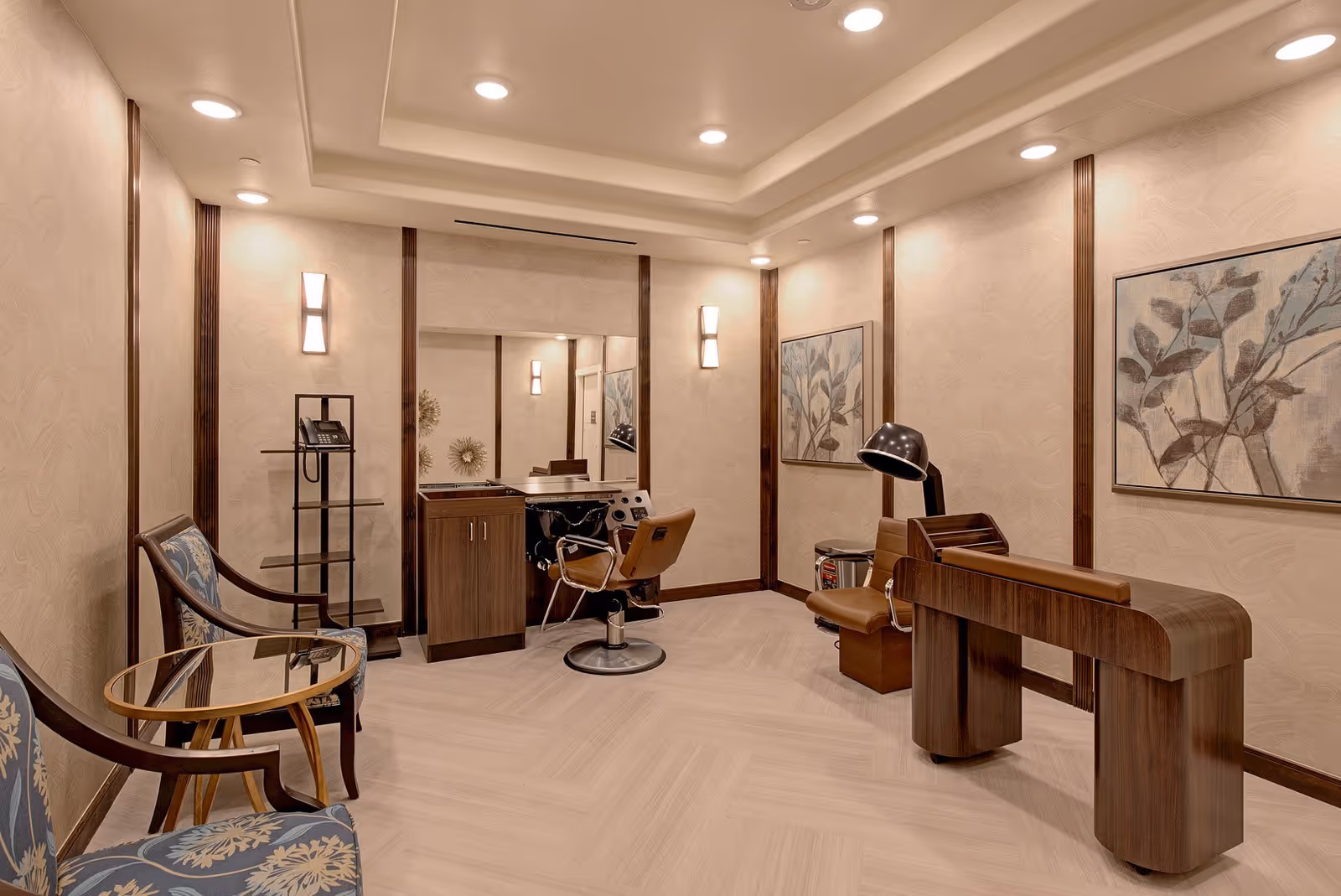 A well-lit salon room in a senior living facility featuring a styling chair in front of a large mirror, a hair dryer chair, two blue patterned armchairs with a small glass-top table between them, and a wooden manicure table. The walls are decorated with framed botanical artwork and have vertical wooden trim accents.