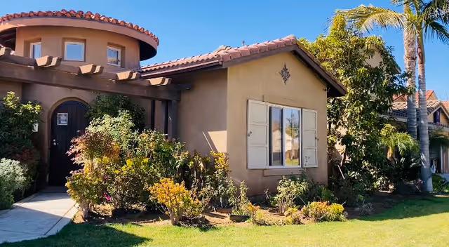 Exterior view of a single-story residential building with a tiled roof, beige stucco walls, and a small garden with various plants and shrubs in front. There is a curved entrance with a wooden pergola and a dark wooden door. Palm trees and other greenery surround the building under a clear blue sky.