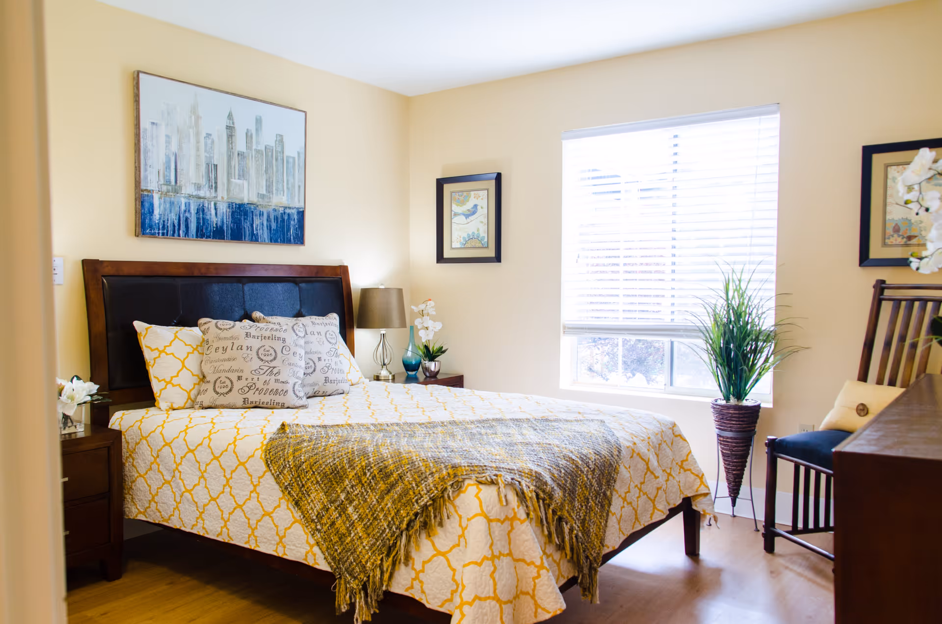 A bright bedroom with a wooden bed frame and a yellow and white patterned bedspread. The bed has decorative pillows and a gray throw blanket. There are two nightstands with a lamp and decorative items, a large window with blinds letting in natural light, a potted plant, a wooden chair with a cushion, and framed artwork on the walls.