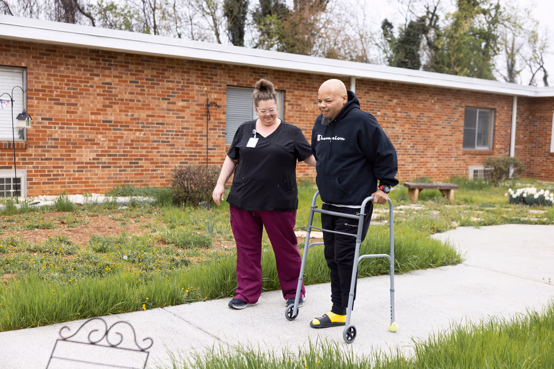 A healthcare worker assists a man using a walker as they walk on a paved path outside a brick building with grass and plants around.