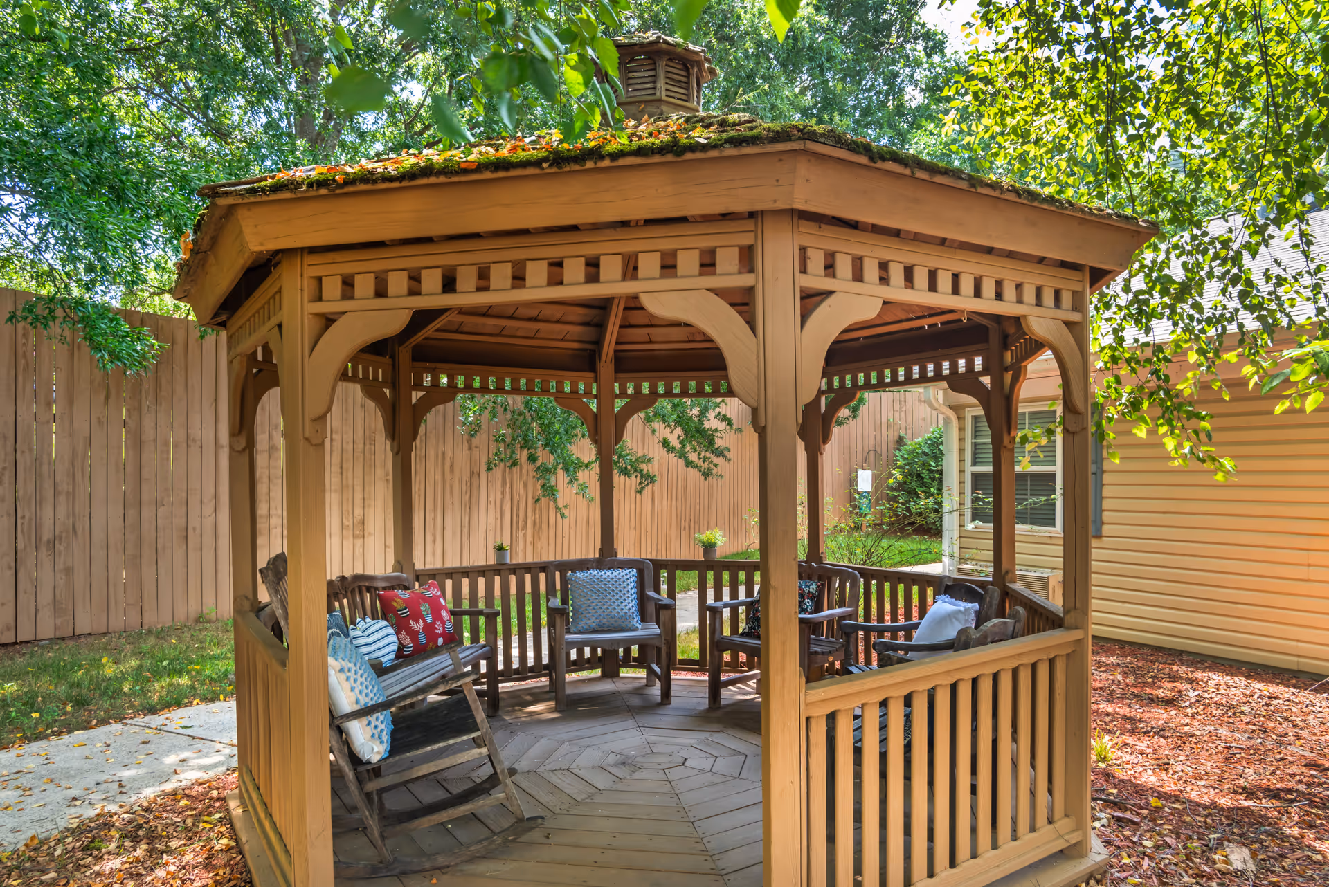 Wooden octagonal gazebo with chairs and colorful cushions set in a shaded backyard.