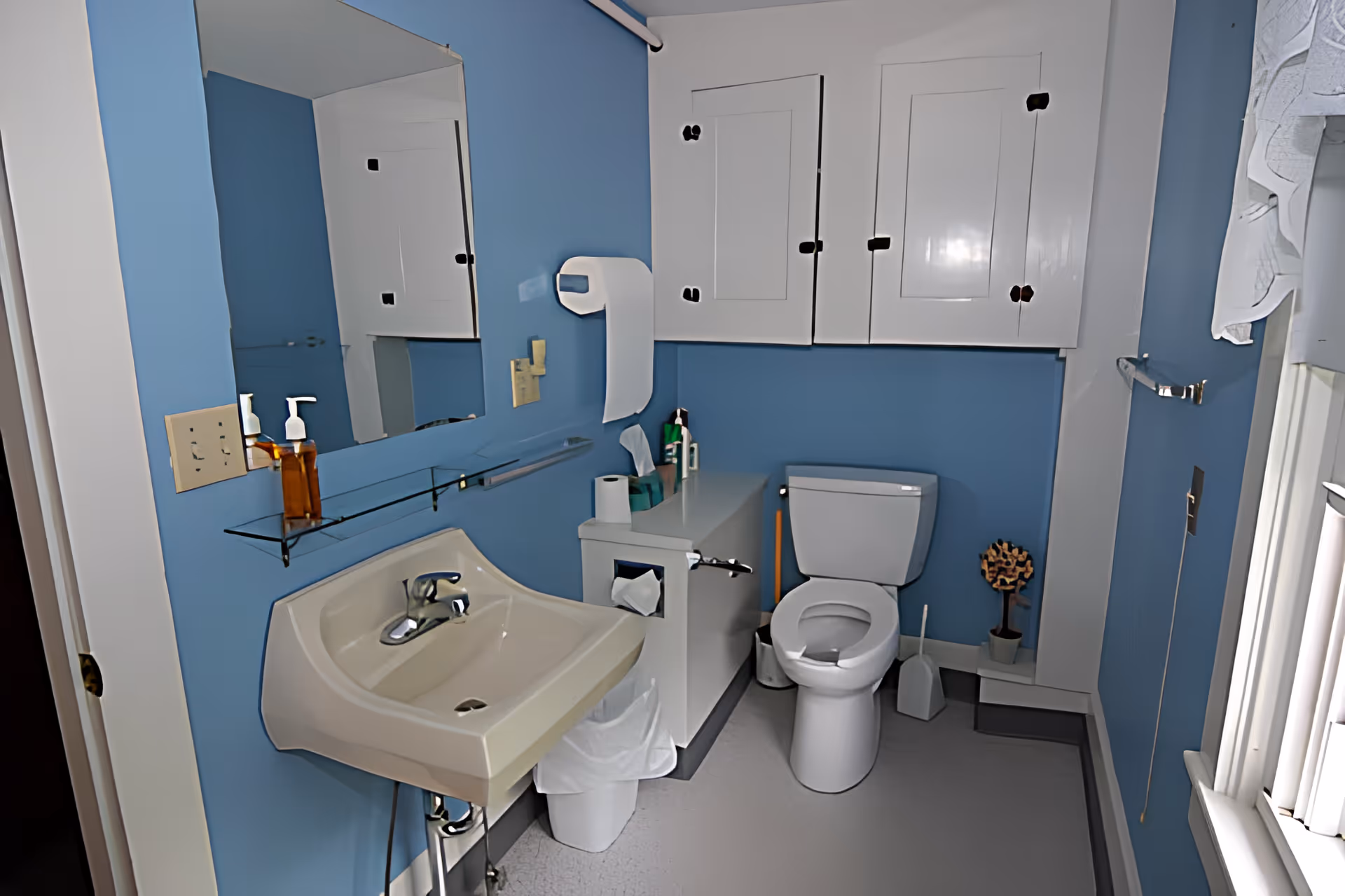 A clean bathroom with blue walls featuring a white sink with a soap dispenser, a large mirror above it, a white toilet, white cabinets mounted on the wall, a tissue box, toilet paper holder, and a small decorative plant near the toilet. There is a window with white blinds on the right side.