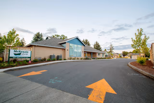 Exterior view of McKenzie Living Eugene facility showing a single-story building with a peaked roof, large windows, and a sign with the facility name and address. The paved driveway has large yellow directional arrows painted on it, and there are trees and landscaping along the sides.