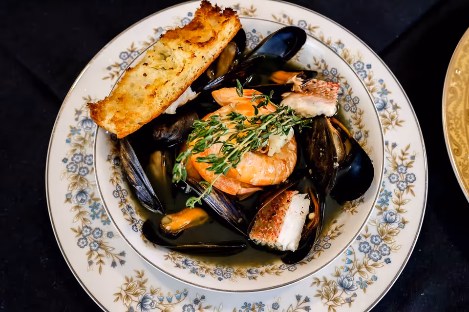 A bowl of seafood stew containing mussels, shrimp, and pieces of fish, garnished with fresh herbs and served with a slice of toasted bread on the side. The bowl is placed on a floral-patterned plate on a dark tablecloth.
