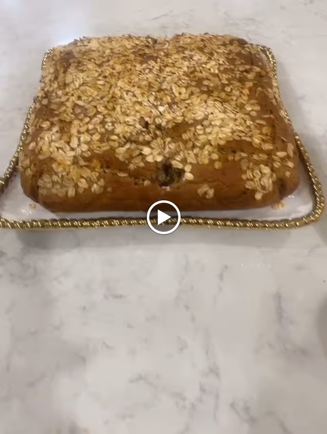 A rectangular baked oatmeal cake topped with rolled oats on a decorative tray with a gold-colored edge, placed on a marble countertop.