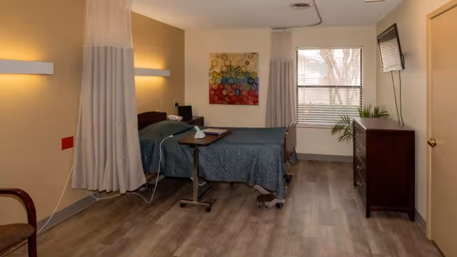 A furnished senior room with a single hospital-style bed, privacy curtain, dresser, wall-mounted TV, and a window.