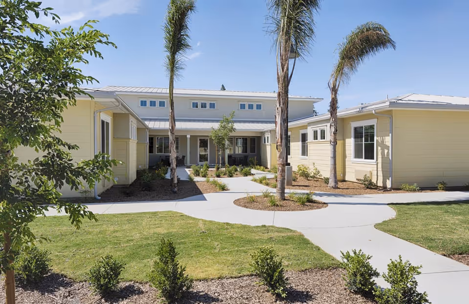 Exterior view of a single-story senior living facility building with light yellow walls, a metal roof, and several windows. The building surrounds a landscaped courtyard with palm trees, small shrubs, and a concrete walkway under a clear blue sky.