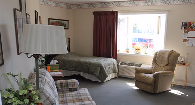 A cozy bedroom with a single bed covered in a green quilt, a plaid sofa, a beige recliner chair, a window with burgundy curtains, and a small table with a lamp and some books. The room has light-colored walls with a floral border near the ceiling.