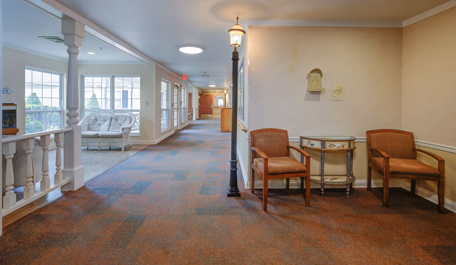 A well-lit hallway in a senior living facility with a patterned carpet floor. On the right side, there are two wooden chairs with cushions and a small decorative table between them. A vintage-style lamp post stands next to the chairs. On the left side, there is a white railing and a seating area with a white cushioned loveseat near large windows letting in natural light. The hallway extends towards a reception desk in the background.