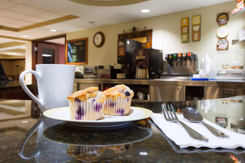 Two blueberry muffins on a plate with a coffee mug and utensils on a counter in a cafe-style dining area with beverage machines in the background.