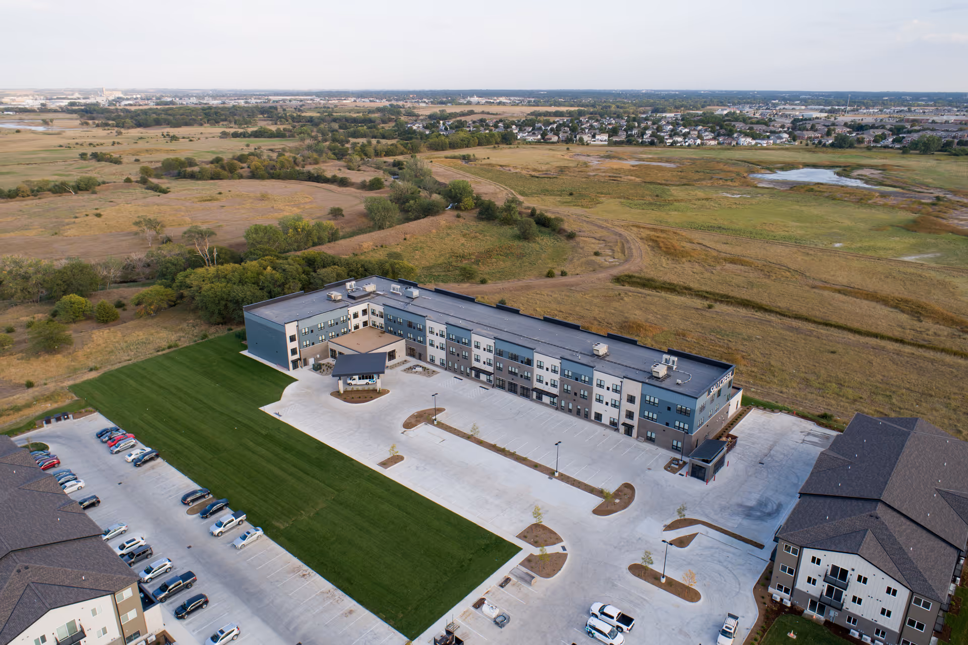 Aerial view of Salt Creek Senior Living facility showing a large, modern three-story building surrounded by parking lots and green lawns, with open fields and a distant residential area in the background.