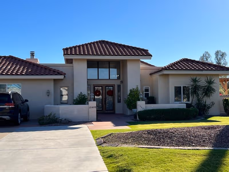 Front exterior view of a single-story building with a tiled roof, beige walls, and a double glass door entrance. There are two small walls with address numbers 10405 and 10401 on either side of the entrance. The driveway and well-maintained lawn with some shrubs and trees are visible under a clear blue sky.