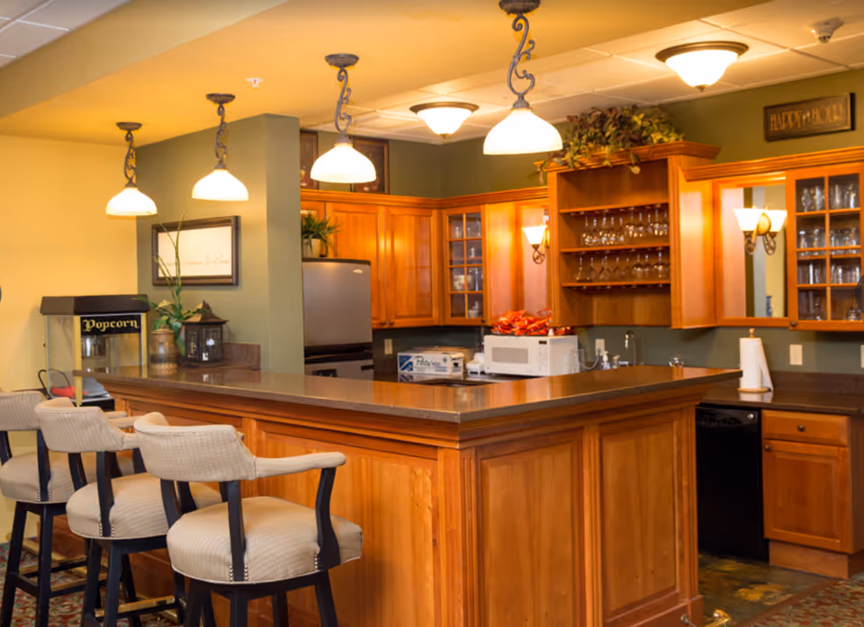 A warm and inviting kitchen area with wooden cabinets, a countertop bar with three cushioned bar stools, hanging pendant lights, a popcorn machine, a microwave, and glassware displayed on shelves.