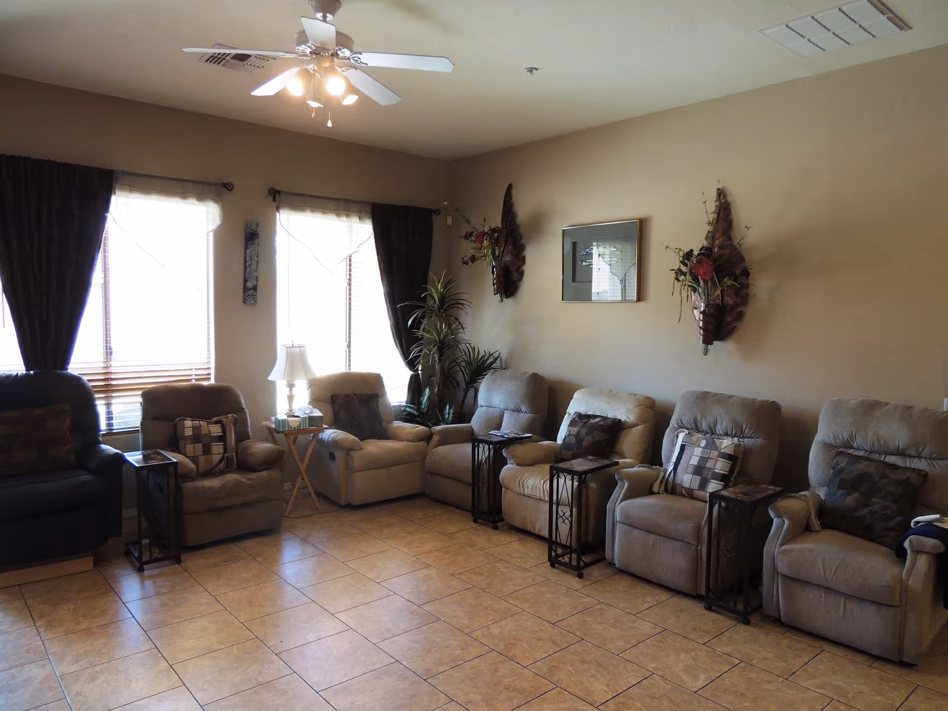 A cozy living room with six recliner chairs arranged in a row along a beige wall. Each chair has a decorative pillow and a small side table next to it. Two large windows with dark curtains and sheer white blinds allow natural light to fill the room. The floor is tiled, and the ceiling has a white fan with lights. Wall decorations include two floral wall sconces and a framed picture. There is also a tall potted plant in the corner.