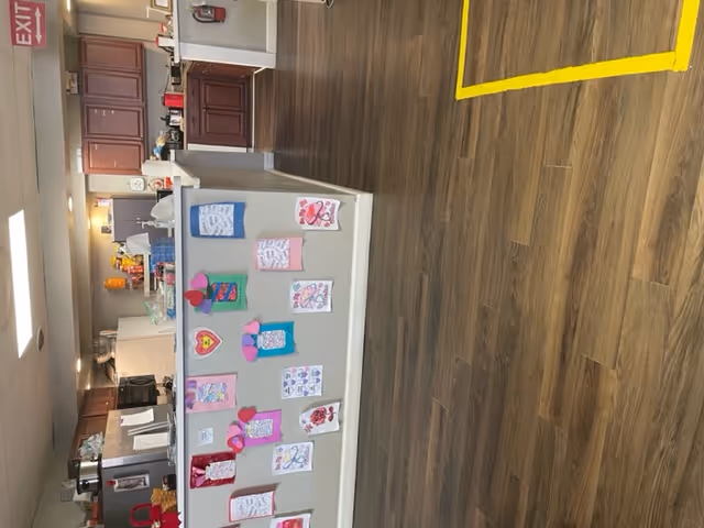 Interior view of a kitchen area in a personal care home with wooden cabinets, a refrigerator, microwave, and various kitchen items on the counters. The front of the kitchen counter is decorated with colorful handmade drawings and crafts. The floor is wood-patterned, and there is a yellow tape marking on the floor. An exit sign is visible on the ceiling.