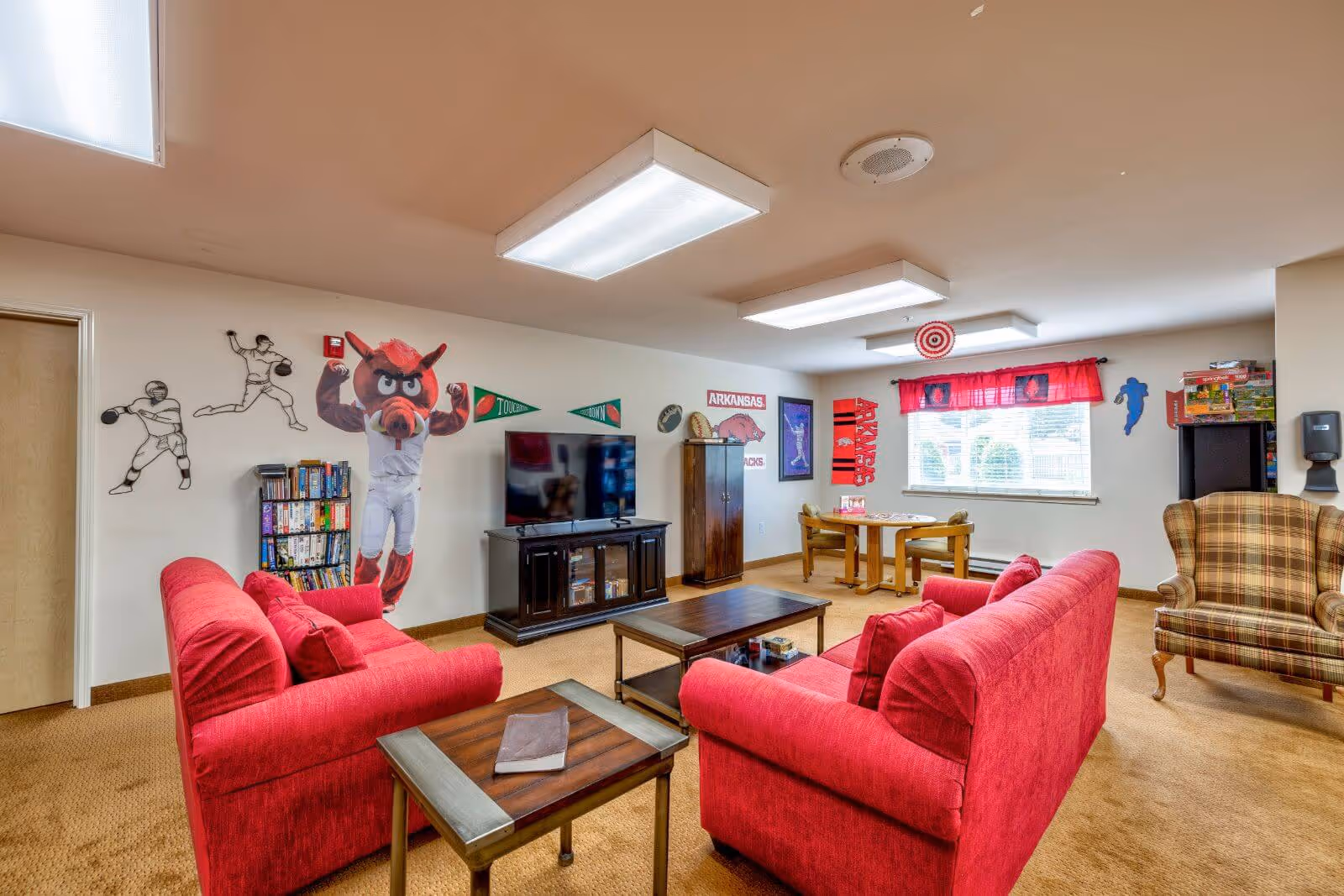 A cozy living room with two red sofas facing a flat-screen TV on a dark wooden stand. The room has sports-themed wall decorations including a large mascot figure, pennants, and sports player decals. There is a wooden coffee table and side table, a plaid armchair, and a small table with chairs near a window with red curtains. The carpet is beige and the ceiling has rectangular fluorescent lights.
