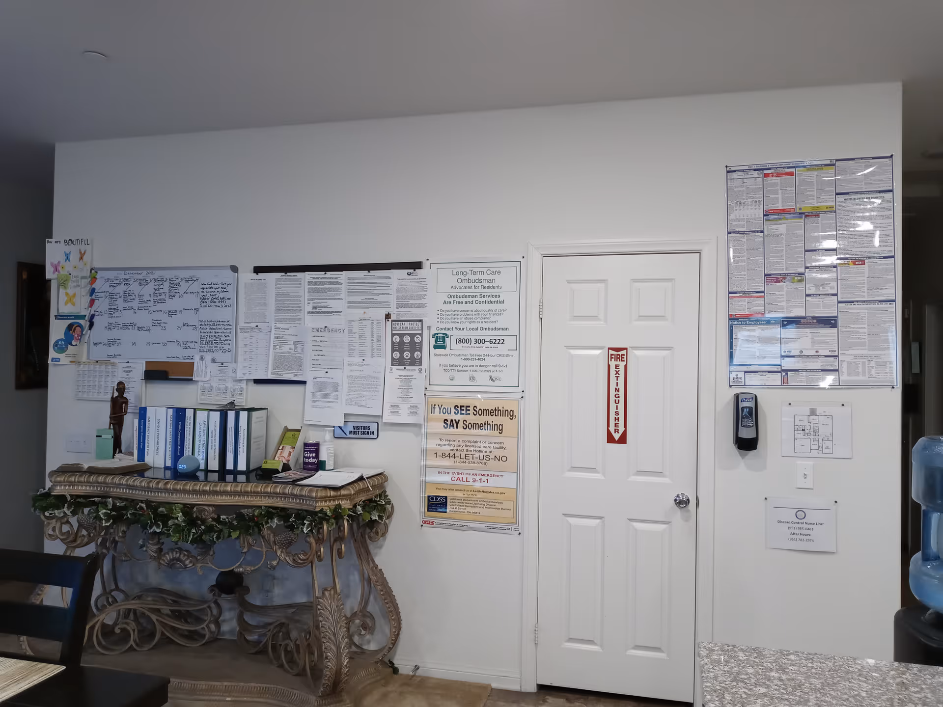 Interior view of a senior home care facility showing a white wall with multiple notices, schedules, and informational posters pinned on it. There is a white door with a fire extinguisher sign, a hand sanitizer dispenser mounted on the wall, a decorative table with binders and a small statue, and a water cooler partially visible on the right side.