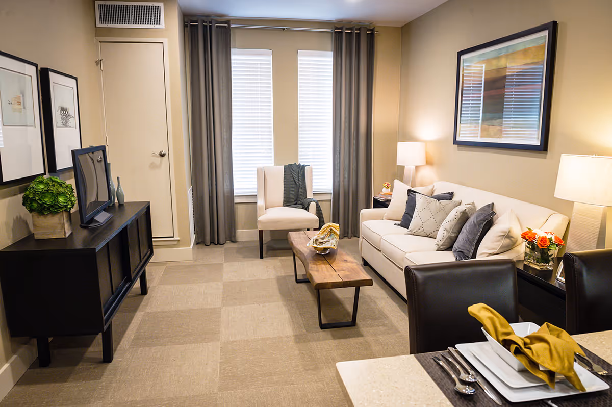 A cozy living room in a senior living facility featuring a white sofa with multiple cushions, a wooden coffee table, a single armchair, two table lamps, framed artwork on the walls, and a TV on a black console. The room has beige walls, checkered carpet flooring, and two windows with gray curtains.