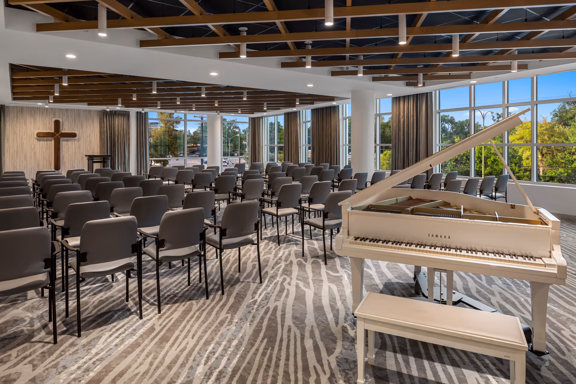 A spacious room with rows of gray chairs arranged facing a wooden podium and a large wooden cross on the wall. A white Yamaha grand piano with a matching bench is positioned in the foreground near large windows that let in natural light and show greenery outside. The ceiling features wooden beams and modern lighting fixtures.