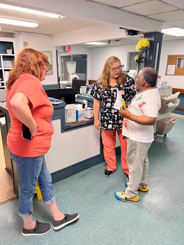 Three people standing and talking inside a healthcare facility reception area. One person is wearing a coral shirt and jeans, another is in medical scrubs with a cat pattern, and the third person is holding papers and wearing a white t-shirt and light pants. The background shows a reception desk with medical supplies and office equipment.