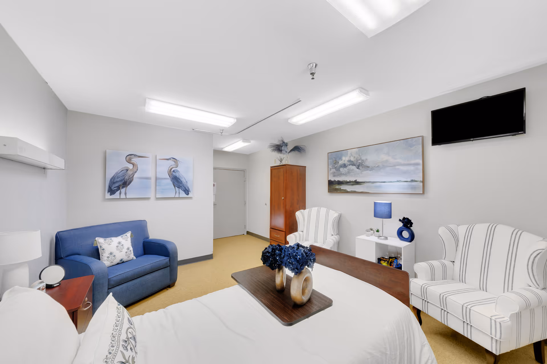 A bright and clean bedroom in a senior living facility featuring a bed with white linens and decorative pillows, a wooden tray with blue flowers on the bed, a blue loveseat with a patterned pillow, two white armchairs with blue stripes, a wooden cabinet, a small white side table with a blue lamp and decorative items, a wall-mounted flat-screen TV, and artwork depicting birds and a landscape on the walls.