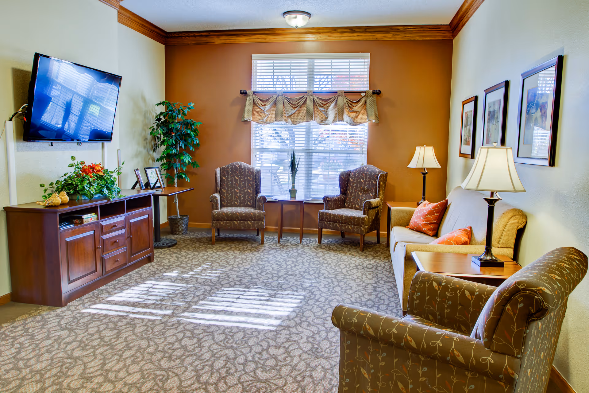 Sunlit communal living room with upholstered chairs and sofa, a wall-mounted TV on a wooden console, table lamps, and a window with a valance.