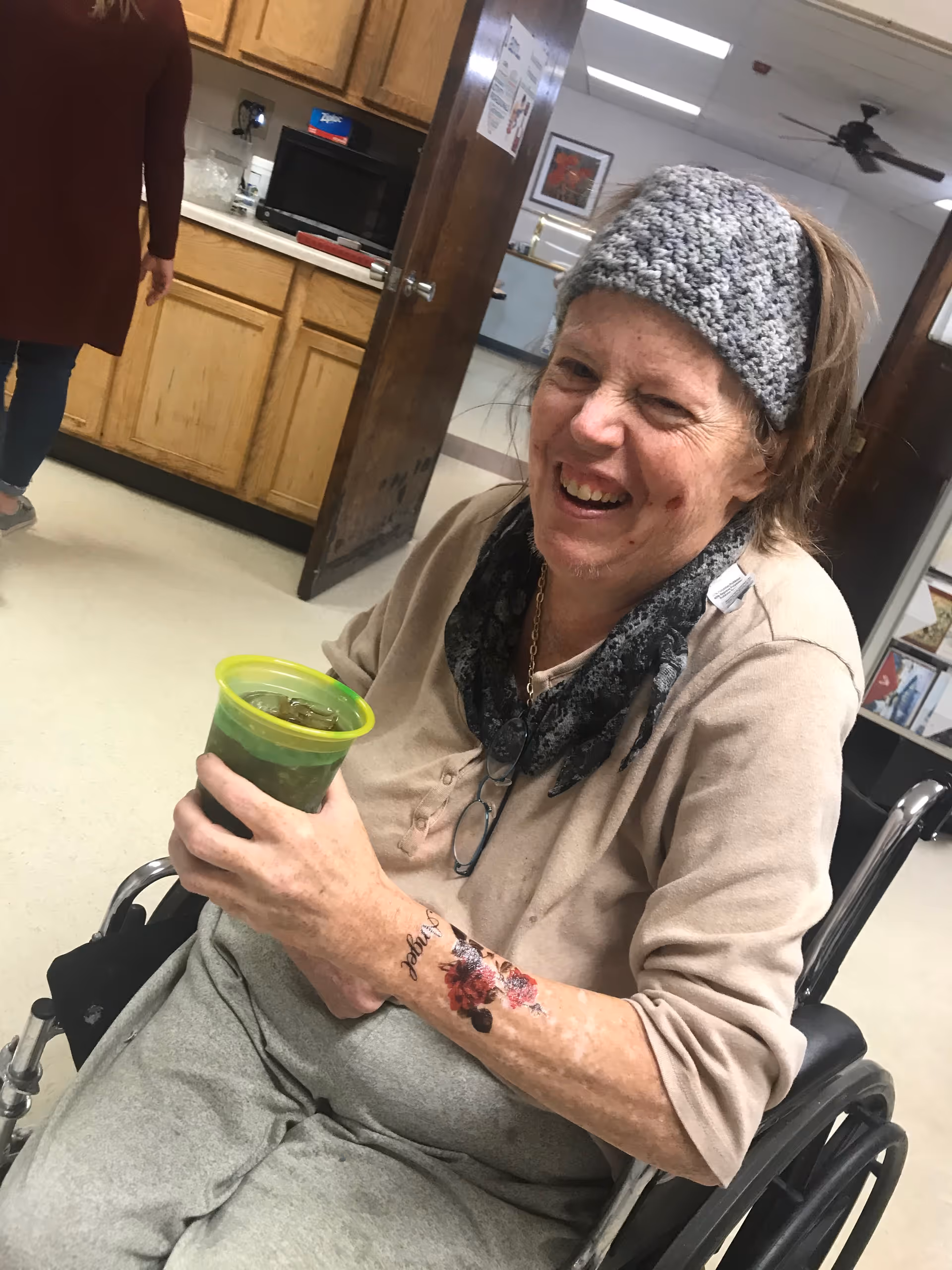 An elderly woman sitting in a wheelchair indoors, smiling and holding a green cup with a drink. She is wearing a gray knitted headband, a beige long-sleeve shirt, gray pants, and a dark scarf. There is a floral tattoo on her forearm. In the background, there is a kitchen area with wooden cabinets and a microwave, and another person walking away.