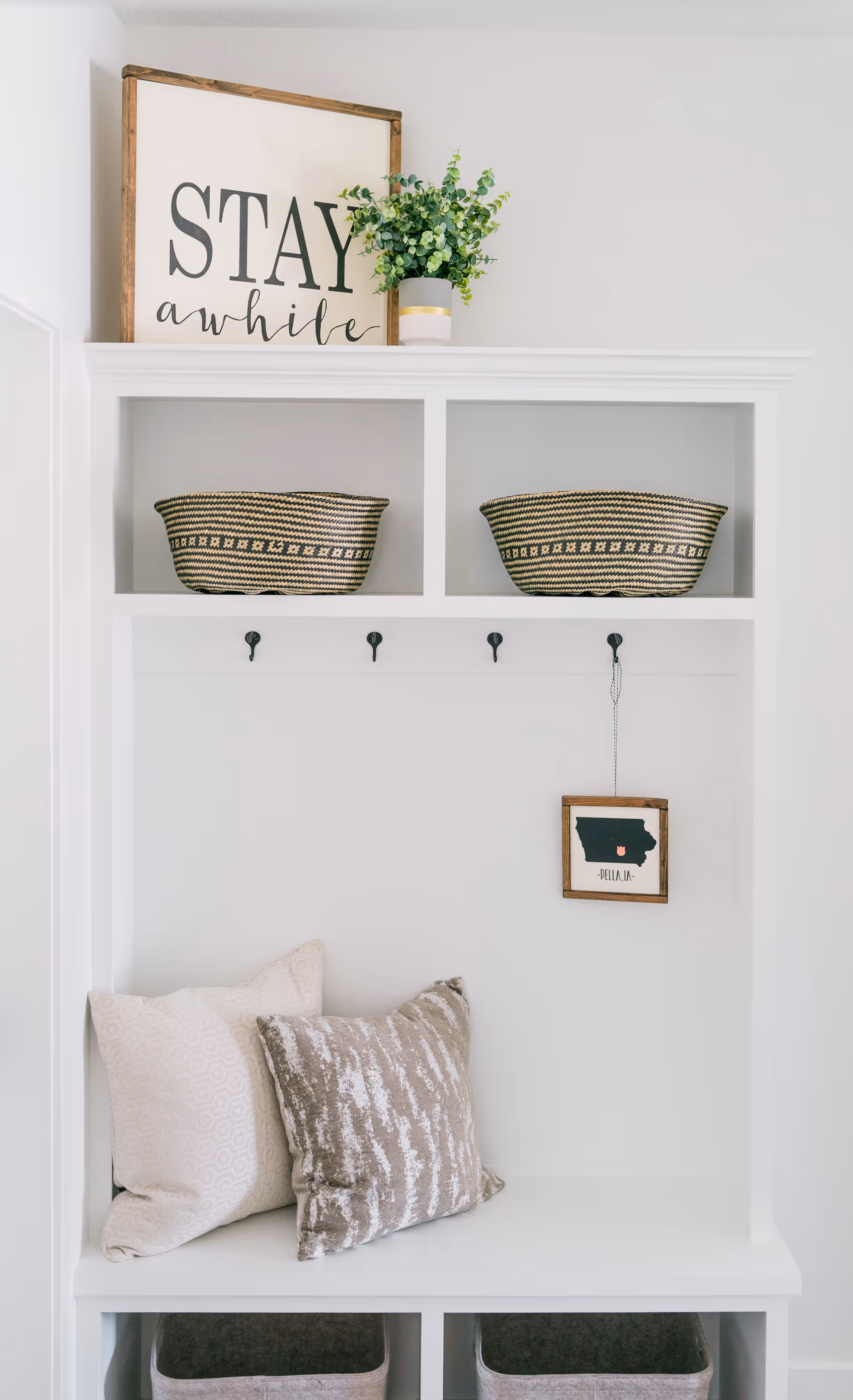 A white built-in bench with two decorative pillows, two woven baskets on the upper shelves, a small potted plant, and a framed sign that says 'STAY awhile'. There are four black hooks on the back panel, with a small framed map of Pella, IA hanging from one of the hooks.