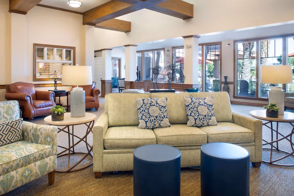 A bright and spacious living room area with a beige sofa adorned with two blue and white patterned pillows, two round blue ottomans in front, a patterned armchair with a decorative pillow, and two side tables each holding a lamp and a small plant. The room features large windows letting in natural light, wooden ceiling beams, and a decorative wire horse sculpture on a table in the background.