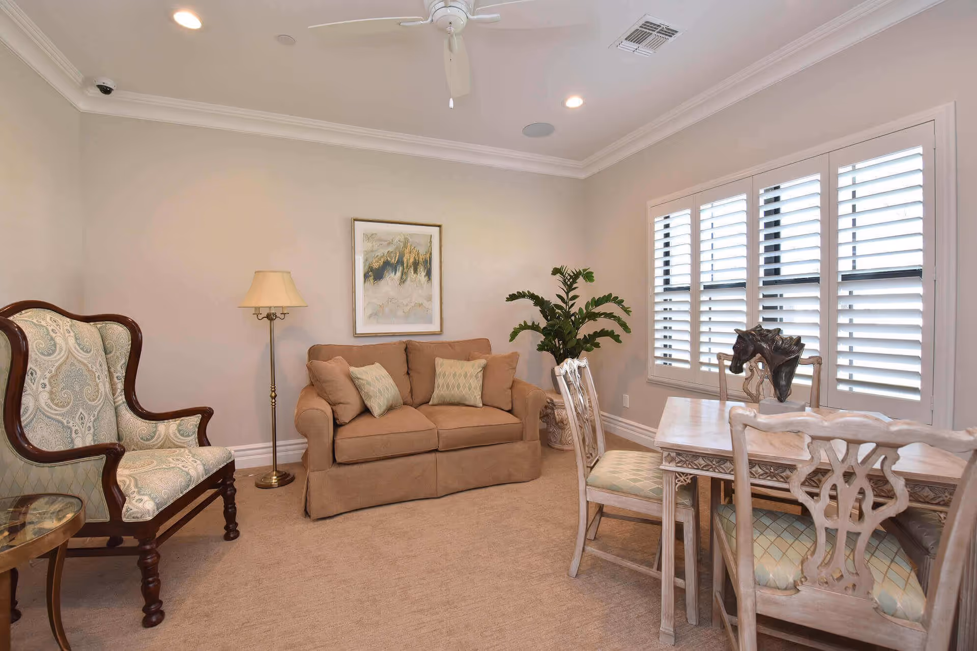A cozy living room with a beige loveseat adorned with two patterned pillows, a vintage-style armchair with intricate upholstery, a floor lamp, a framed abstract painting on the wall, a potted plant in the corner, and a wooden dining table with four chairs near a window with white plantation shutters.