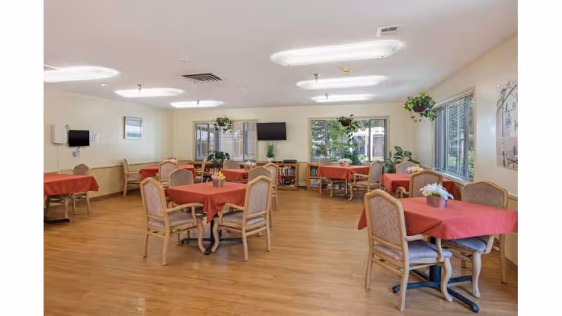 A dining room in a senior living facility with several tables covered in red tablecloths, each surrounded by four cushioned chairs. The room has wooden flooring, large windows with blinds, potted plants on the windowsills, and a television mounted on the far wall. The walls are light-colored, and the ceiling has fluorescent lighting fixtures.
