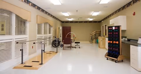 A spacious rehabilitation room with parallel bars for walking exercises, a wheelchair, exercise equipment including colorful hand weights on a rack, and various therapy tools. The room has large windows with blinds, light-colored walls, and bright overhead lighting.