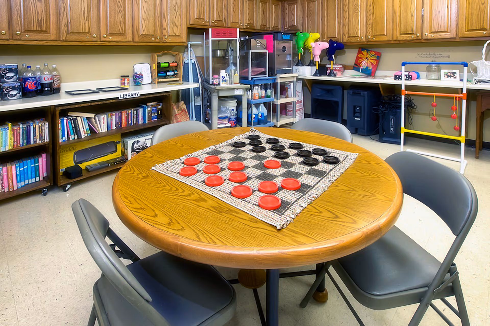 A round wooden table with a checkers game set up on a woven board, surrounded by four gray folding chairs. In the background, there are wooden cabinets above a countertop with various items including soda bottles, bookshelves labeled 'Library' filled with books, and shelves with craft supplies and toys. The room appears to be a recreational or activity room.