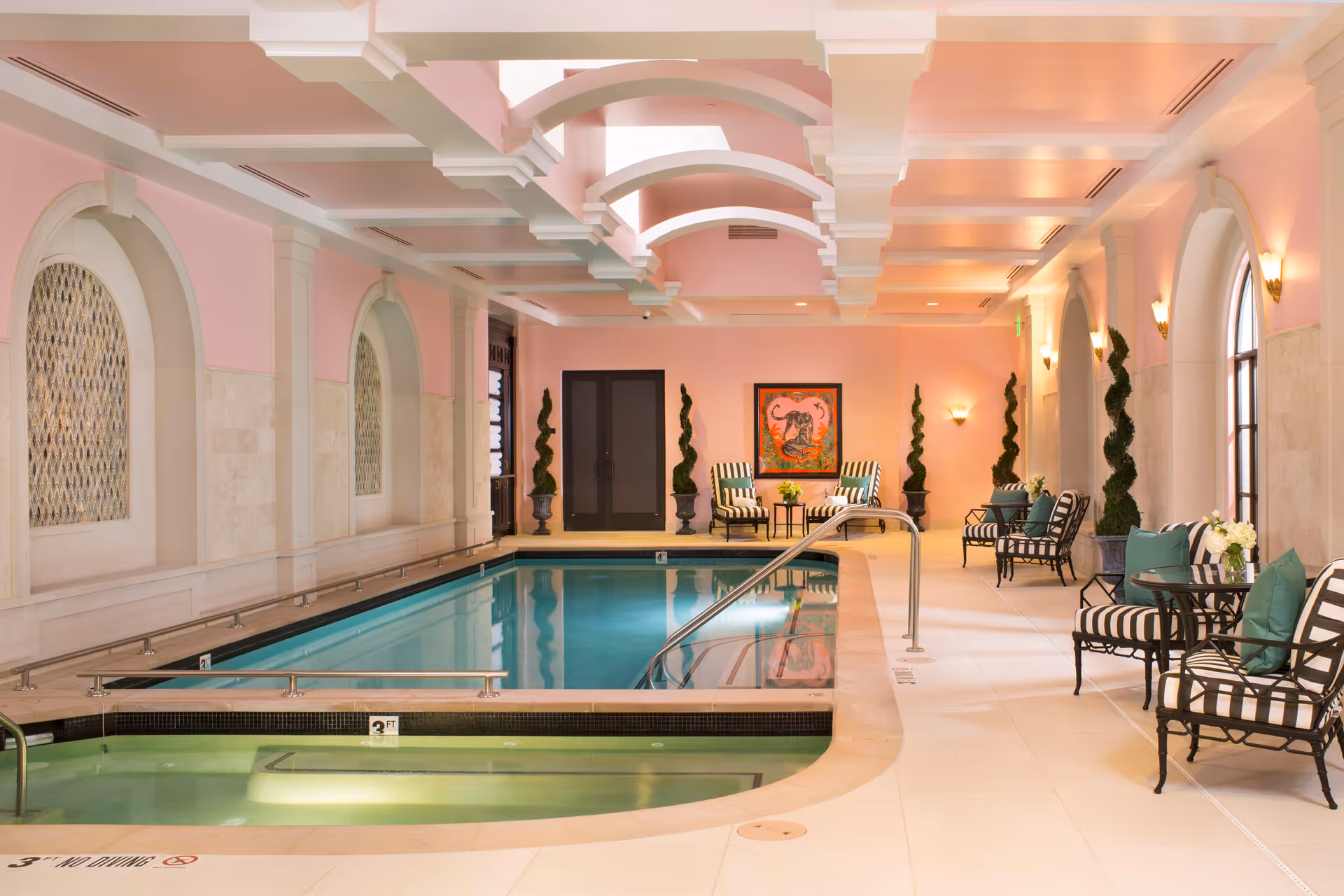 Indoor swimming pool area with a smaller adjacent pool or hot tub, surrounded by cushioned black and white striped chairs with green pillows. The walls are painted pink with arched windows and decorative wall sconces. The ceiling features white beams with skylights, and there are tall potted topiary plants along the walls.