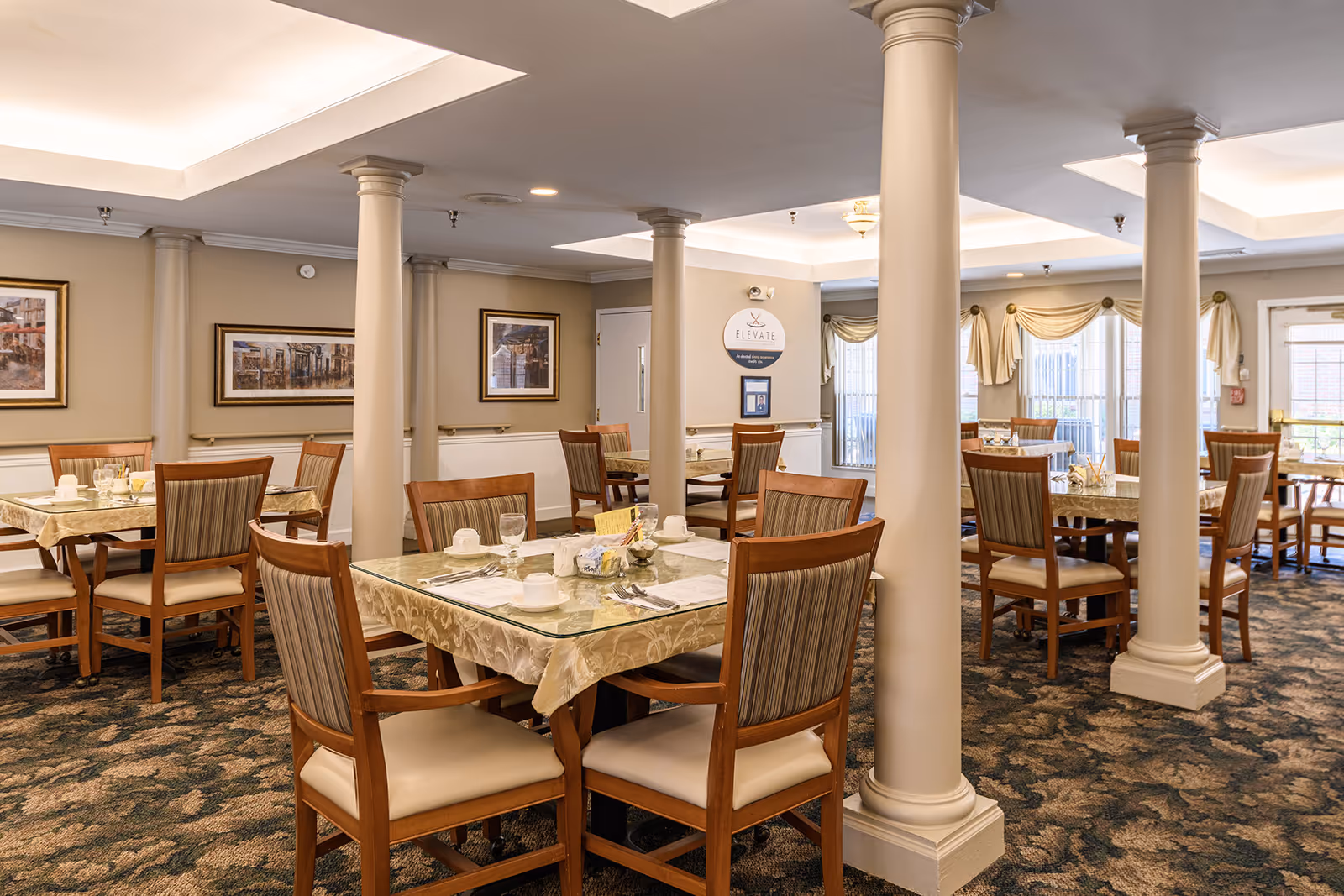 A dining room in a senior living facility with multiple tables covered with beige tablecloths and glass tops. Each table is set with white cups, glasses, and silverware. The room features beige walls, decorative columns, framed artwork, and large windows with draped curtains allowing natural light to enter.