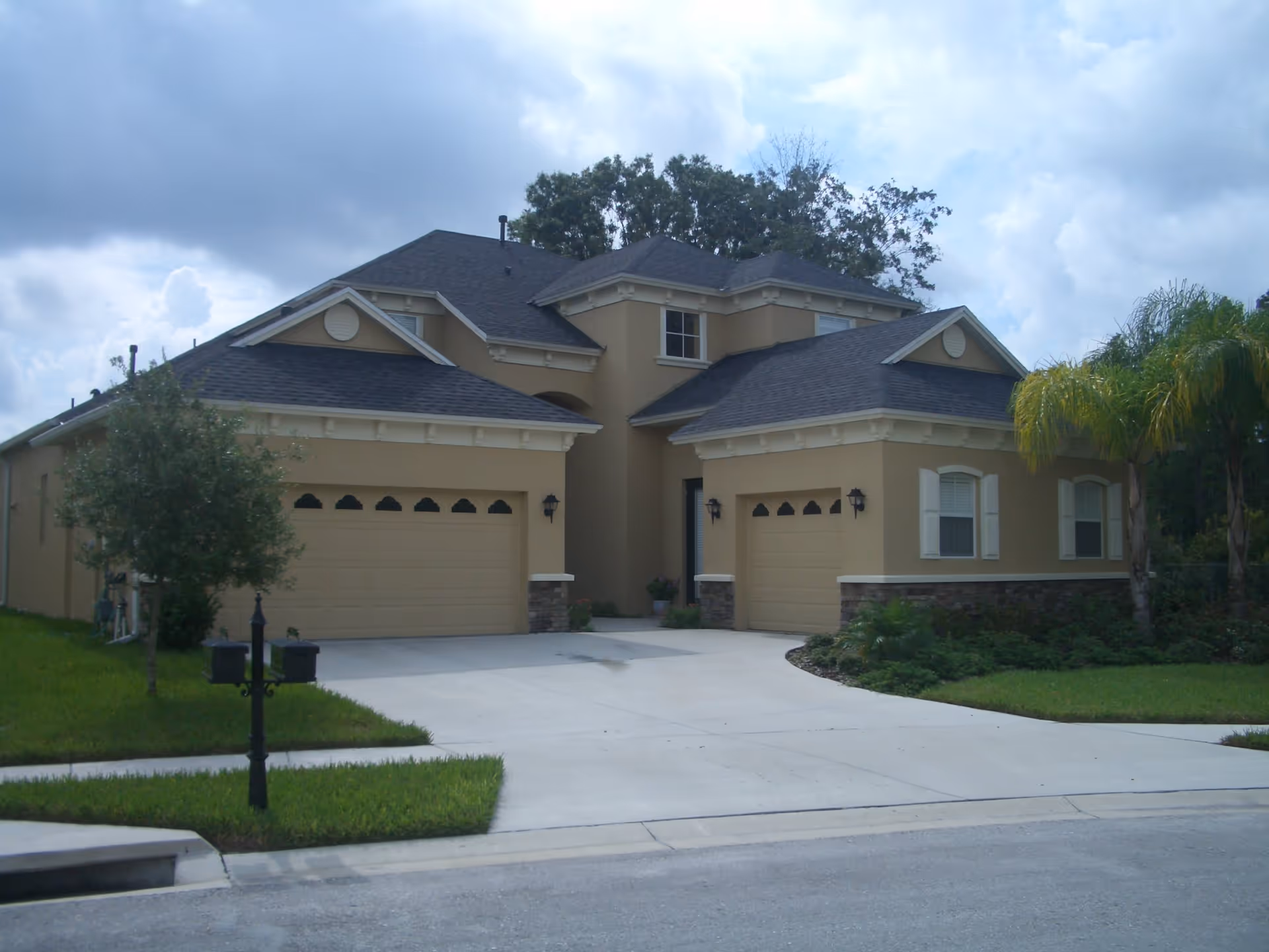 Two-story beige suburban house with attached garages, a wide driveway, mailbox and palm tree under a cloudy sky.