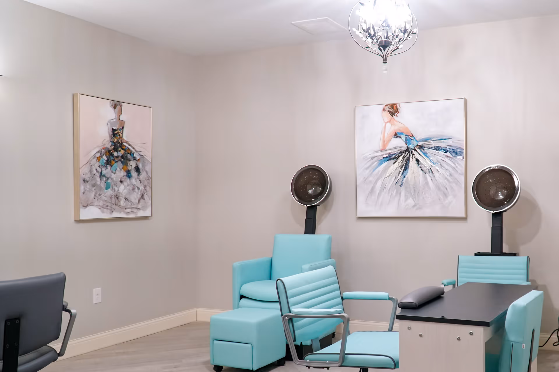 Interior of a salon area with light blue salon chairs, two hair dryer stations, and two paintings of women in dresses on the walls. The room has light-colored walls and flooring, with a decorative ceiling light fixture.