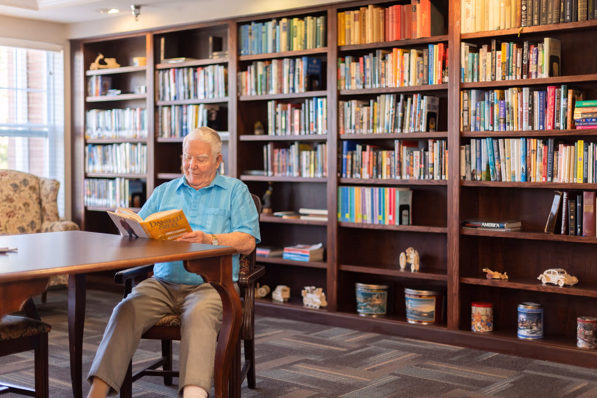 An elderly man with white hair wearing a blue shirt and gray pants is sitting at a wooden table reading a Danielle Steel book in a room with large wooden bookshelves filled with books and decorative items. There is a floral armchair near a window with blinds in the background.