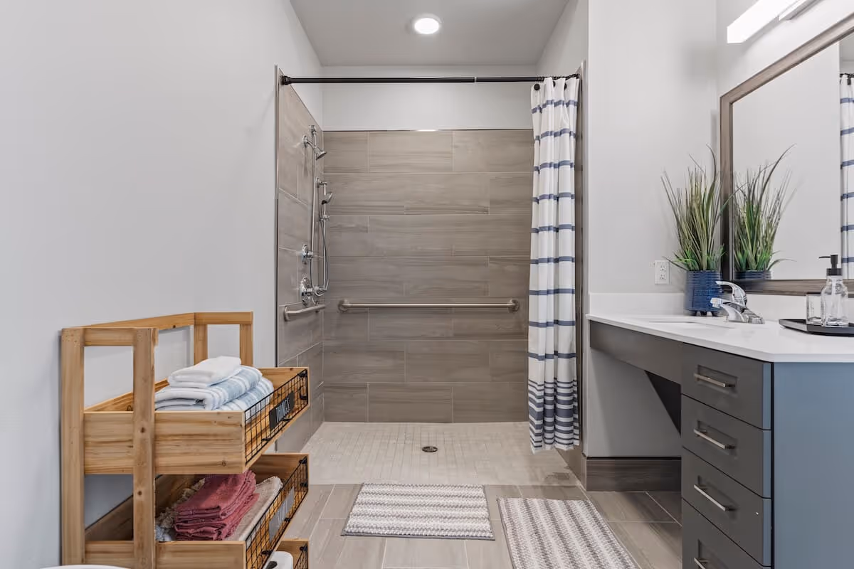 A modern bathroom featuring a walk-in shower with gray tiles and a striped shower curtain. There is a wooden shelf unit holding folded towels on the left side. On the right, there is a vanity with a sink, a large mirror, and two potted plants. The floor has two striped rugs.