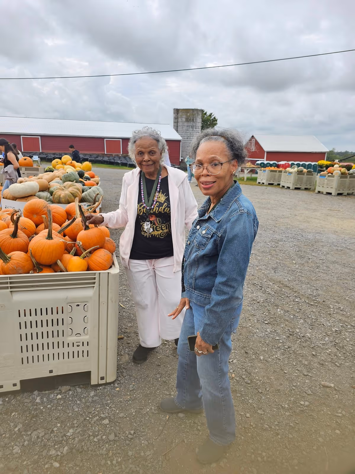 Two elderly women standing outdoors at a pumpkin patch with bins full of pumpkins and gourds. One woman is wearing a white jacket and pants with a black shirt, and the other is wearing a denim jacket and jeans. Red barns and a cloudy sky are visible in the background.