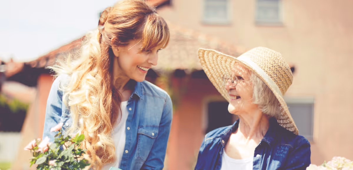 A younger woman with long blonde hair and a denim shirt smiling and holding flowers while talking to an elderly woman wearing glasses and a large straw hat, both outdoors with a building in the background.